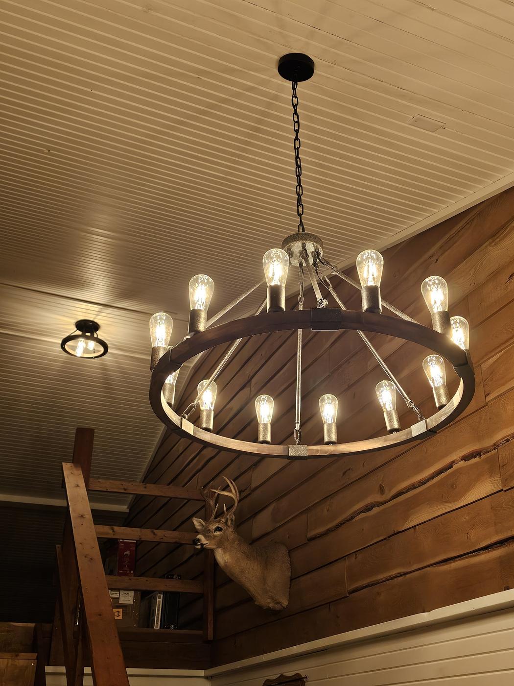 Round wagon wheel chandelier with exposed bulbs hung from a cabin ceiling